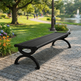 A black outdoor park bench with slatted seating and curved legs, placed on a stone pathway in a landscaped park with grass, trees, flowers, and a pond in the background.