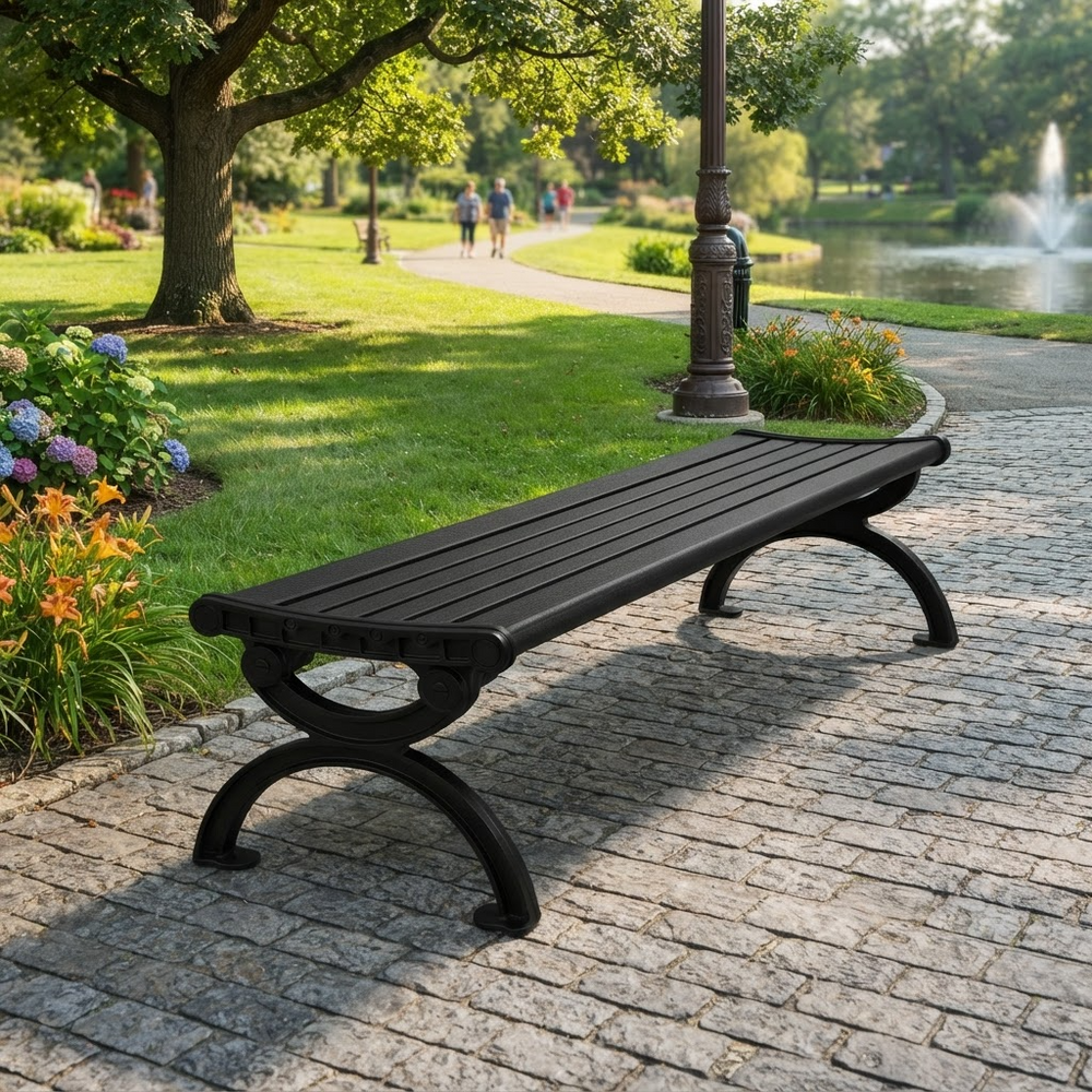A black outdoor park bench with slatted seating and curved legs, placed on a stone pathway in a landscaped park with grass, trees, flowers, and a pond in the background.