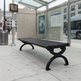 A black outdoor bench with ornate curved metal legs sits on a city sidewalk under a glass-covered bus stop shelter. A transit schedule board, trash can, and city street scene are visible in the background.