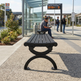 A black outdoor bench with a slatted top and curved metal base, placed on a paved station platform. A person sits on the bench in the background near a bus stop shelter, with other people and station signage visible.