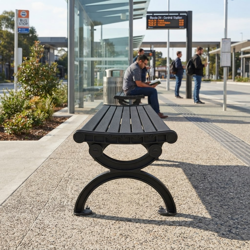A black outdoor bench with a slatted top and curved metal base, placed on a paved station platform. A person sits on the bench in the background near a bus stop shelter, with other people and station signage visible.