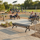 A modern black metal park bench in the foreground of a sunny playground, with several people sitting on benches in the background on a paved walkway.