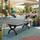 A black slatted park bench with decorative curved legs sits on a colorful playground surface in a real outdoor play area. Children and adults are seated nearby under beige canopy tents, with playground equipment, trees, and potted plants in the background.