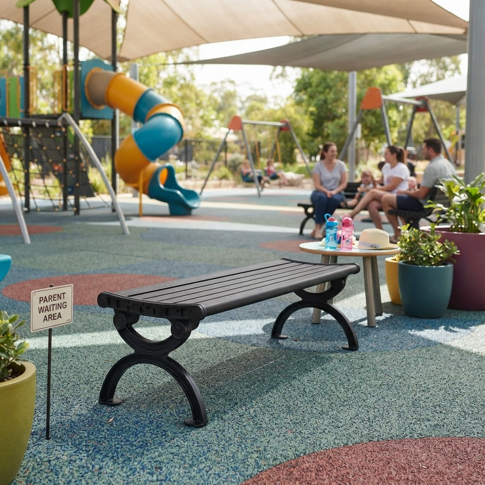 A black slatted park bench with decorative curved legs sits on a colorful playground surface in a real outdoor play area. Children and adults are seated nearby under beige canopy tents, with playground equipment, trees, and potted plants in the background.