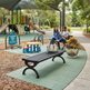 A woman and man sit on a black park bench in a playground setting, with a smaller black bench in the foreground holding a tote bag and water bottles. The scene includes a children's play area, shade structure, trees, and a green turf surface.