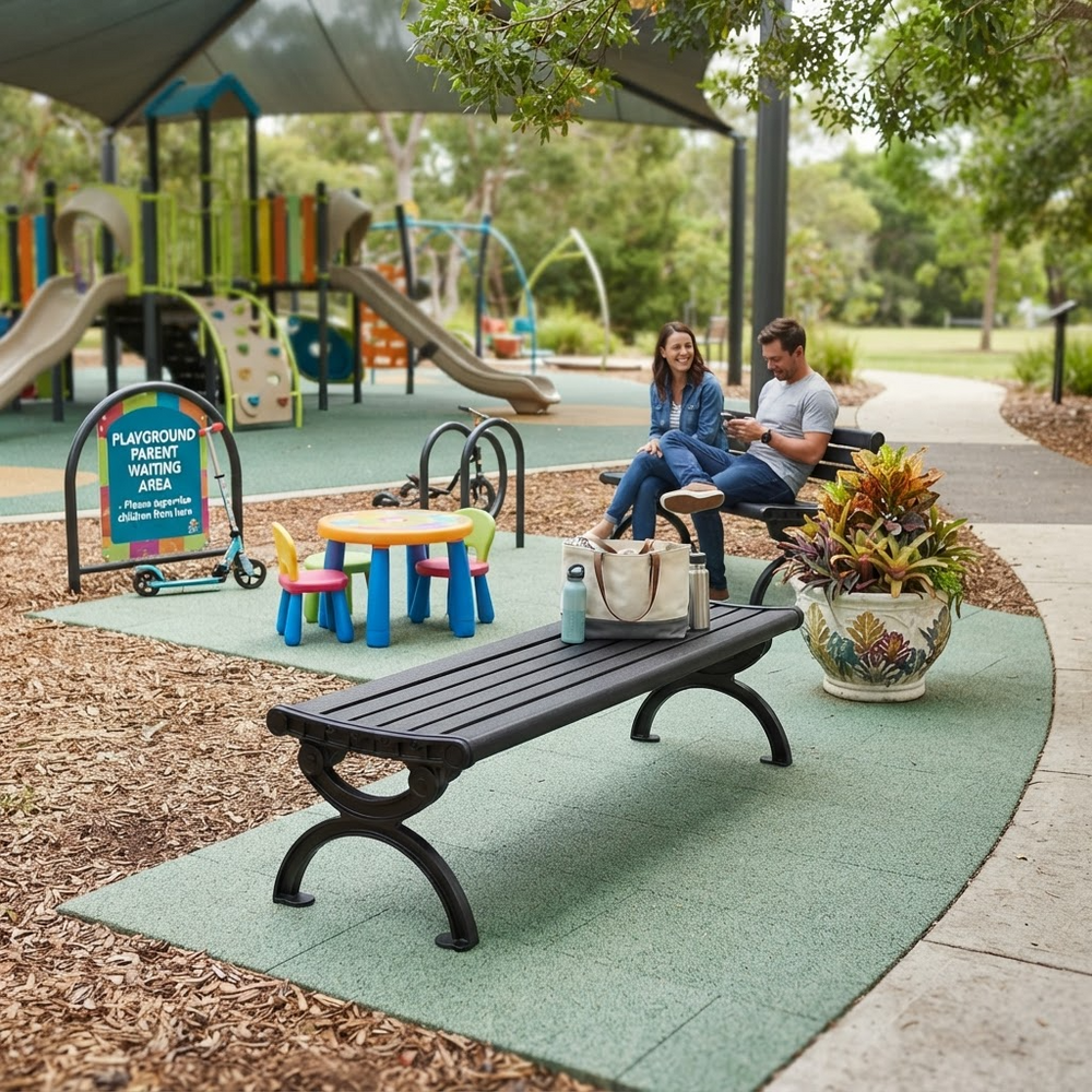 A woman and man sit on a black park bench in a playground setting, with a smaller black bench in the foreground holding a tote bag and water bottles. The scene includes a children's play area, shade structure, trees, and a green turf surface.