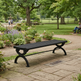 A black metal park bench with decorative curved legs, situated on a stone pathway in a lush green park with flower beds and trees.