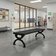 A black outdoor-style bench with curved legs is placed in a modern indoor hallway. Large glass windows look into an indoor basketball court where people are playing.