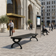 A modern black outdoor bench and matching kiosk stand in a city plaza in front of a grand stone building, with several people walking and sitting nearby.