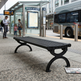 A black metal public transit bench in a city station area, with people standing at a bus stop and a bus visible in the background.