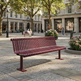 A modern slatted park bench in a deep burgundy red, situated in a paved urban plaza with trees, historic buildings, and a few seated people in the background.
