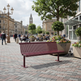 A modern maroon slatted public bench in a paved city plaza, surrounded by large planters with colorful flowers, trees, and pedestrians in the background.