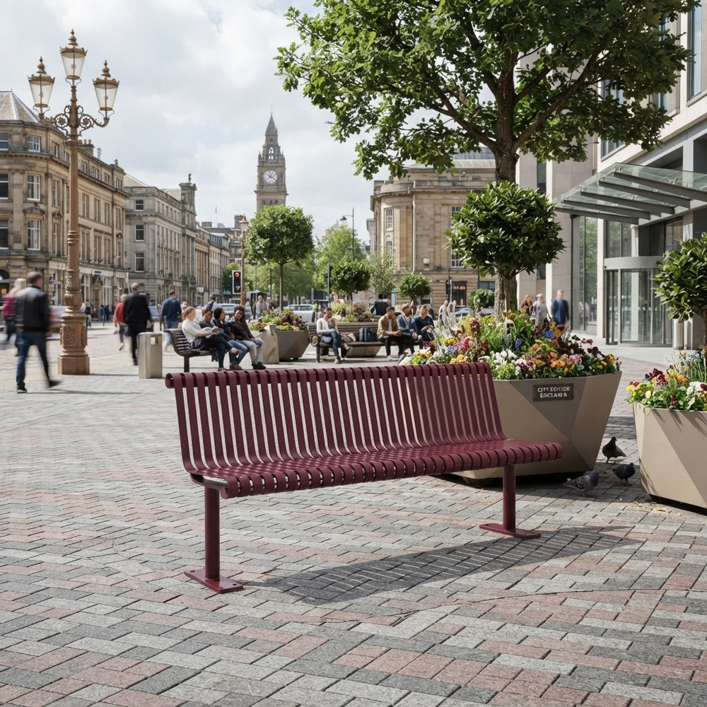 A modern maroon slatted public bench in a paved city plaza, surrounded by large planters with colorful flowers, trees, and pedestrians in the background.