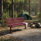 A reddish-brown slatted outdoor bench sits on a gravel campsite path in a forest, with a green tent and fire pit visible in the background.