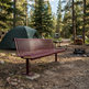 A red-brown slatted metal campsite bench in a forest clearing, with a green tent, picnic table, fire pit, and trees in the background.