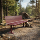 A red metal park bench in a forest campsite setting, with trees, a tent, and a campfire visible in the background.