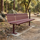 A metal park bench with a reddish-brown slatted seat and back, installed on a concrete pad in a wooded campsite setting with trees, rocks, and a tent in the background.