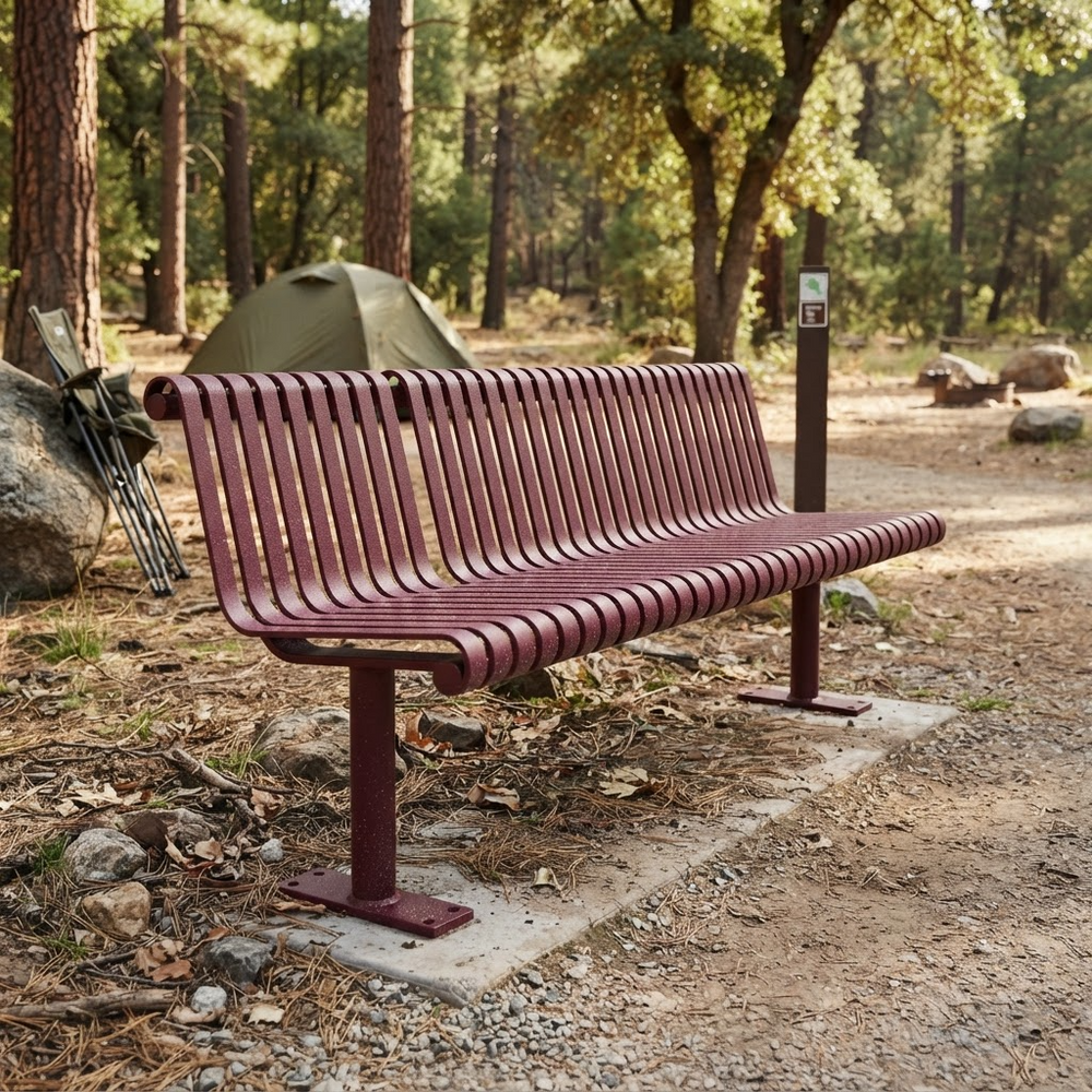 A metal park bench with a reddish-brown slatted seat and back, installed on a concrete pad in a wooded campsite setting with trees, rocks, and a tent in the background.