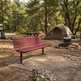 A red wooden park bench in the foreground at a forest campsite, with a beige tent, picnic table, fire pit, and trees in the background.