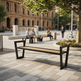 A modern black metal bench with a light wood seat is shown in an outdoor urban plaza with people sitting and walking in the background.