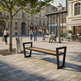 A modern outdoor bench with a black metal frame and a light wood seat is shown in a real urban plaza with people sitting and walking, buildings in the background, and a tree nearby.