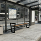 A modern outdoor bus stop bench with a black metal frame and brown wooden seat, situated under a glass shelter at a city transit station. A city bus and street scene are visible in the background.