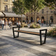 A modern outdoor bench with a black metal frame and a brown wooden seat is situated in a paved public square with people walking and trees in the background.