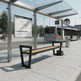 A modern outdoor bus stop shelter with a bench, glass panels, and a transit map display, shown in a real urban environment with a bus and street in the background.