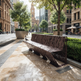 A modern sculptural metal bench with a laser-cut leaf pattern in a city plaza, with trees, planters, and a street scene in the background.