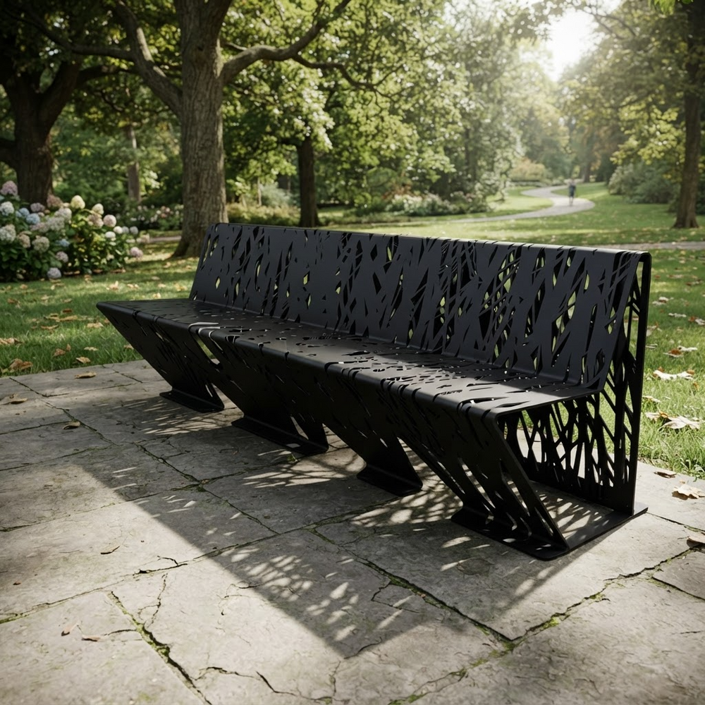 A modern black metal park bench with a leaf-like cutout pattern, set on a stone patio in a lush green park.