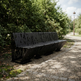 A modern black metal park bench with a leaf-cutout pattern, situated on a gravel path next to lush green trees.