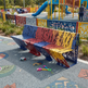 A colorful metal bench with a tree-bark cutout pattern, painted in blue, red, and yellow sections, situated in a children's playground with kids and slides in the background.
