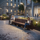 A modern urban plaza at dusk with a sculptural dark metal bench, two people sitting on one end, and a woman reading on the bench. The scene includes lit street lamps, stone buildings, planters, and a paved walkway.