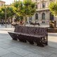 A modern dark brown metal bench with an intricate laser-cut lattice pattern, situated in a sunny, tree-lined public square with people sitting on other benches in the background.