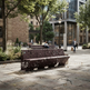 A modern outdoor urban bench with a dark brown perforated metal frame and matching seat, situated on a paved plaza in a city setting with trees, buildings, and pedestrians visible.