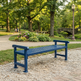 A dark blue slatted outdoor park bench placed on a gravel path in a sunny, tree-filled park with a stone bridge and lamppost in the background.