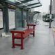 A bright red wooden bus stop bench sits on a wet gray concrete platform at a city bus station, with a bus shelter, maps, bicycles, and a green bus in the background.