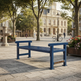 A blue wooden park bench in a paved public square with trees, a historic building, and a few distant people in the background.