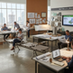 A modern office interior with several people working at desks, some seated on long bench-style chairs. The space includes large windows, architectural drawings on tables, lamps, and a wall display labeled 'Materials Library' with sample panels and a screen.