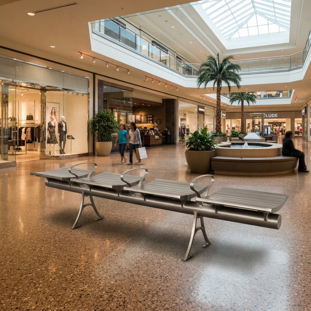 A modern stainless steel outdoor-style bench with curved armrests sits on a polished terrazzo floor in a large, sunlit shopping mall atrium with other seating, plants, and storefronts visible in the background.