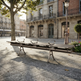 A long outdoor bench with five seats and integrated armrests, shown in a sunlit urban plaza with stone buildings, trees, and a lamppost in the background.