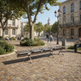 A modern metal bench with integrated cup holders is shown in a real outdoor plaza with cobblestone ground, trees, benches, people walking, and historic buildings in the background.