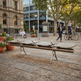 A modern outdoor bench with a brushed metal frame and light wood slats, situated in a city plaza with people, trees, and buildings in the background.