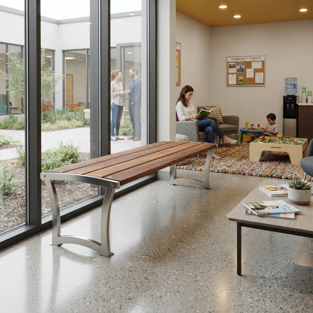 A modern outdoor-style bench with a brown wooden slatted seat and curved silver metal legs, placed in a bright office lobby next to large floor-to-ceiling windows. A woman sits on a nearby sofa reading, and a child plays on a small table in the background.
