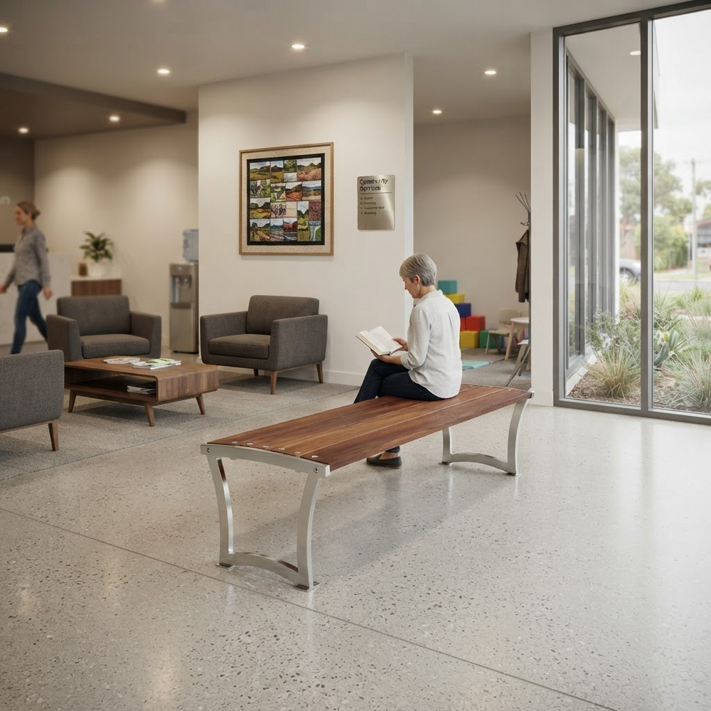 A woman sits on a wooden bench with metal legs in a modern office lobby, reading a book. The space includes gray upholstered chairs, a coffee table, framed wall art, and large floor-to-ceiling windows.