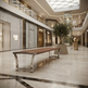 A modern wooden bench with curved stainless steel legs situated in a luxury, upscale shopping mall corridor with visible Dior, Gucci, and Cartier storefronts.