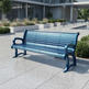 A modern outdoor bench with blue metal slats and armrests, positioned on a paved plaza in front of a contemporary building with greenery.