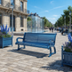 A bright blue metal park bench in an outdoor urban plaza with stone paving, classical buildings, street lamps, a glass water feature, and people walking in the background.