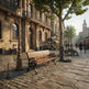 A scenic urban street with ornate stone buildings, cobblestone pavement, a wooden park bench in the foreground, lampposts, trees, and a few pedestrians. No product is isolated or being sold; this is a real environment scene, so the best fit is Lifestyle rather than Product/Packaging or Scenery.