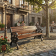 A wooden park bench with ornate black metal arms and legs sits on a cobblestone plaza in a sunlit town square. Stone buildings, a lamppost, a tree, and a few seated people are visible in the background.